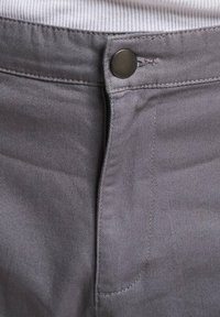 Sive traperice s patent-zatvaračem, okruglom metalnom dugmetu i ravnim šavovima. Tekstura površine s blagim sjajem koja ističe detalje tkanine.