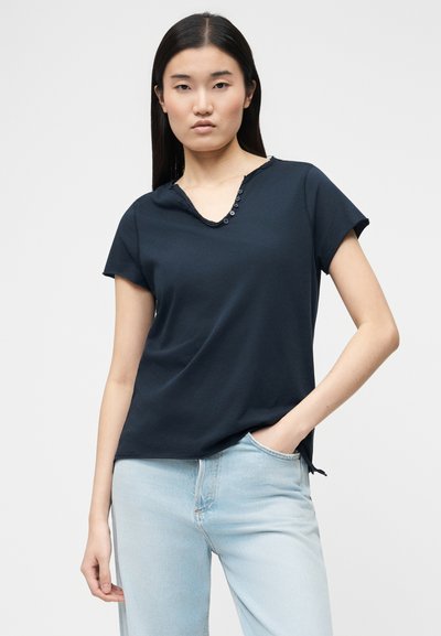 Γυναίκα με μακριά μαύρα μαλλιά που φοράει μαύρο κοντομάνικο μπλουζάκι με V λαιμόκοψη και κουμπιά, και ανοιχτόχρωμο τζιν, ποζάροντας με το ένα χέρι στην τσέπη.