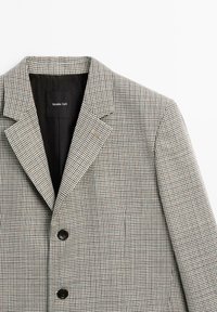 Blazer met een pied-de-poule-patroon in beige en zwart, voorzien van revers met inkepingen en twee knopen aan de voorkant. Label zichtbaar aan de binnenkant van de kraag.