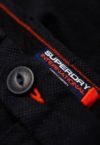 Tessuto nero con trama testurizzata; presenta un'etichetta blu con "SUPERDRY INTERNATIONAL" in bianco e cuciture arancioni; include un bottone grigio.