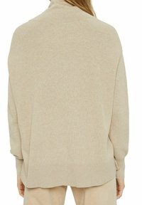 Kvinna iklädd en beige ribbad stickad tröja med nedhasade axlar och långa ärmar, visad bakifrån.