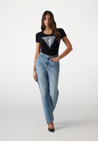 Sort kortærmet t-shirt med et trekantet grafik og teksten "GUESS", parret med lyseblå høj talje jeans og sorte hæle.