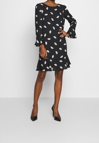 Svart klänning med långa, utsvängda ärmar och en volangnederdel. Har ett vitt lövmönster över hela klänningen och en rund halsringning.