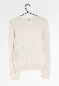 Beige långärmad tröja med rund halsringning, med dekorativa silverdekorationer på vänster sida och ribbade ärmslut och nederkant.