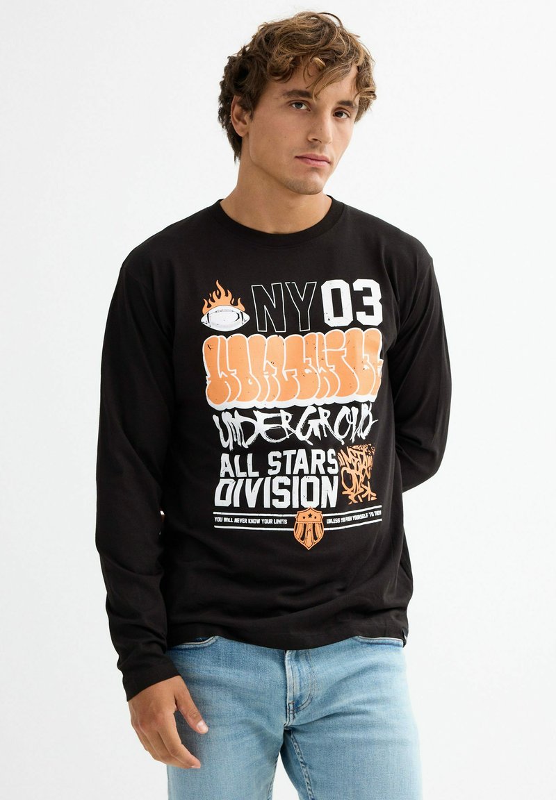 Camisa negra de manga larga con estampados gráficos que incluyen "NY03," llamas y varios textos en naranja y blanco en la parte frontal. Corte casual.