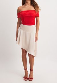 Rød top med bare skuldre og rynket struktur, parret med en lys beige asymmetrisk nederdel. Omfatter røde sandaler med remme og blomsterdetaljer.