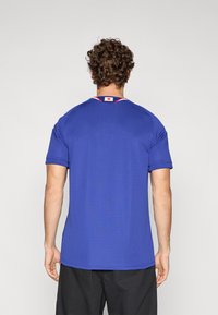 Μπλε αθλητική μπλούζα με κοντά μανίκια, στρογγυλή λαιμόκοψη και κόκκινες λεπτομέρειες. Διαθέτει λογότυπο στο πίσω μέρος του γιακά. Λεπτή υφή.