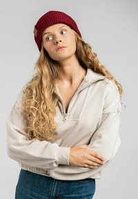 Beżowa bluza rozpinana z rypsowym kołnierzem, połączona z głębokoczerwoną dzianinową czapką. Modelka ma długie, kręcone włosy i ma na sobie niebieskie dżinsy.