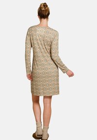 Woman with hair in a bun wearing a knee-length, long-sleeve beige dress with a multicolored floral pattern, beige socks, and brown slippers, facing away.