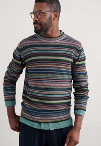 Suéter a rayas multicolores con rayas horizontales en verde, rojo y azul. Cuello y puños acanalados. Lleva una camisa de color azul claro debajo.