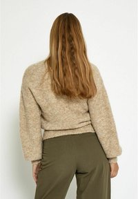 Fuzzy, beige gebreide trui met lange, losse mouwen en een cropped ontwerp. Gecombineerd met olijfgroene broek, gezien van achteren.