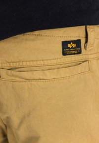 Khaki nohavice vyrobené z odolnej bavlny s jemnou textúrou. Obsahujú zadnú vrecko a čiernu etiketu s oranžovým brandingom.