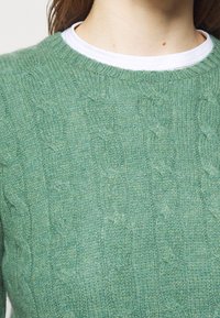 Pull vert en maille avec un motif torsadé texturé, col rond, et sous-shirt blanc superposé visible au niveau du col.