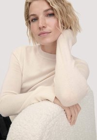 Hellbeige Pullover mit geripptem Ausschnitt und Bündchen. Weicher, strukturierter Stoff. Subjekt hat die Hand auf einer flauschigen, weißen Oberfläche ruhen.