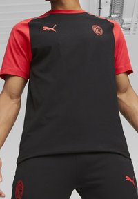 Camiseta deportiva negra con mangas rojas, que presenta un logo de Puma y un emblema del AC Milan. Tejido suave con un diseño ajustado.