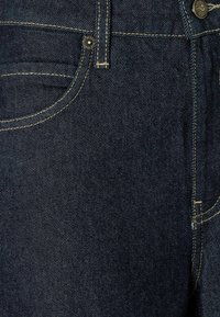 Un jean en denim bleu foncé présente des coutures visibles en jaune, une poche avant et une fermeture à bouton en métal. Le tissu a une texture lisse.