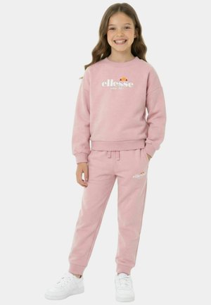 Fille debout et souriante, portant un sweat-shirt rose de la marque ellesse et un pantalon de jogging assorti, avec des baskets blanches sur un fond uni.