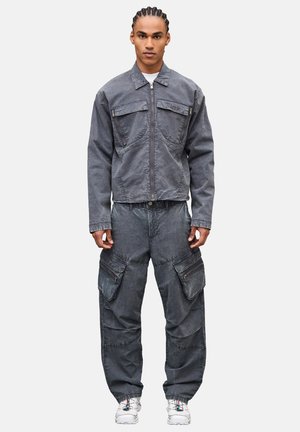 Giovane con capelli intrecciati indossa una giacca grigia con zip e pantaloni cargo con grandi tasche, abbinati a scarpe da ginnastica bianche, in piedi rivolto verso lo spettatore.