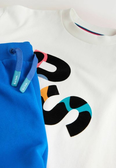 Pantalones deportivos azules con cordones descansando sobre una sudadera blanca que presenta letras grandes, coloridas y parcialmente visibles con estilo.