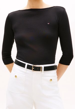 Persoon draagt een nauwsluitende zwart shirt met driekwart mouwen, een klein logo, en een witte broek met hoge taille, gouden knopen op de zakken en een zwarte riem.