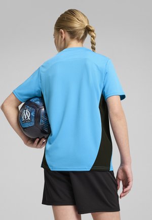 Modré sportovní tričko s krátkým rukávem, černé boční panely a materiál odvádějící vlhkost. Černé šortky a modrý fotbalový míč jsou drženy.