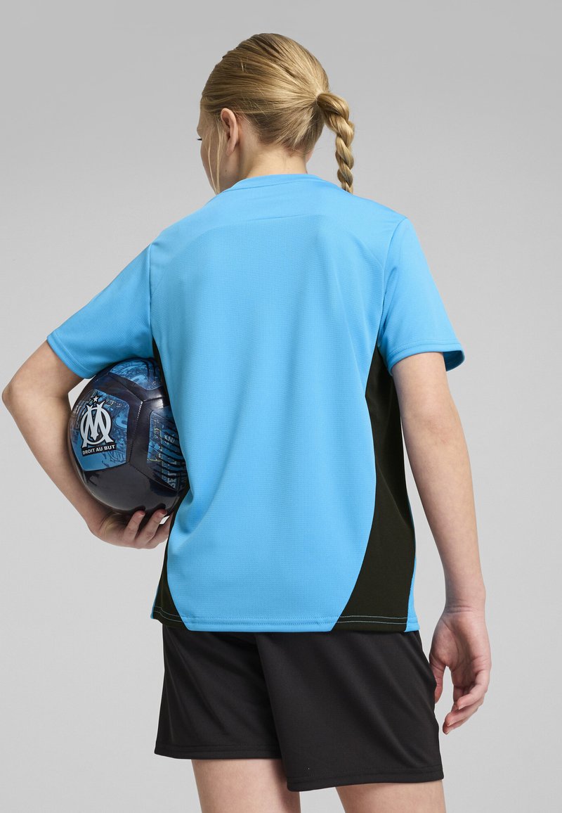 Zila sporta krekls ar īsām piedurknēm, melniem sānu paneļiem un mitrumu izsviedošu audumu. Tur rokās melnas šorti un zila futbola bumba.