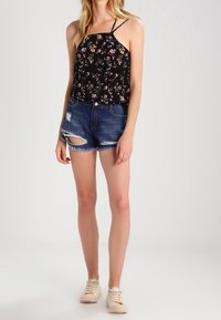 Débardeur noir à fleurs avec des fines bretelles, associé à un short en denim bleu déchiré. Des baskets beiges viennent compléter la tenue.