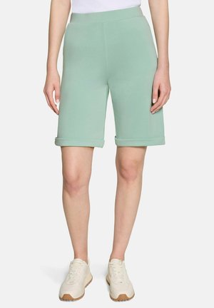 Kobieta w miętowych, krótkich spodenkach do kolan z podwiniętymi brzegami, białych trampkach i włożonej białej koszuli, stojąca na neutralnym tle.