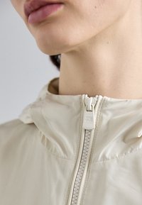 Casaco capucho em bege claro com um colarinho alto e fecho frontal. Material suave com um brilho sutil e detalhe de puxador de fecho com a marca.