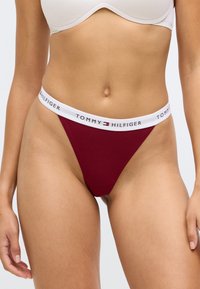 Burgunderfargett truse med en hvit elastisk stropp som har "TOMMY HILFIGER"-logo. Glatt stoff, lav midje-design, minimal dekning på sidene.
