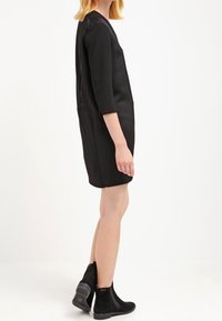 Robe noire à manches courtes avec un design simple et une coupe droite. Portée avec des bottines noires chevauchantes avec une finition texturée et un léger talon.