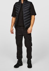 Sort puffervest over en sort T-shirt, kombineret med cargobukser og sorte sneakers. Har lynlåslukning og sidelommer.