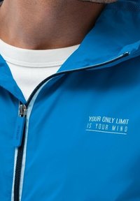 Blå letvægtsjakke med lynlåslukning foran, hætte og trykt tekstdetalje på brystet. Glat stof med et elegant design.