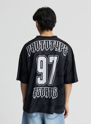 Persona con capelli neri corti che indossa una maglia nera oversize con la scritta "PROTOTYPE 97 REDANDS" in font gotico bianco sulla schiena.