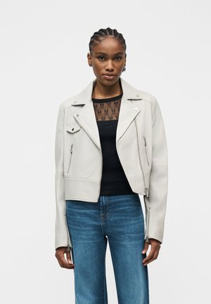 Hajfonatokkal rendelkező nő világosszürke, cipzáras motoros dzsekit visel fekete hálós felső és kék farmer felett, egy egyszínű háttér előtt áll.