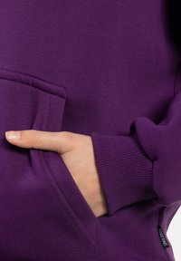 Sudadera morada con un bolsillo frontal, puños acanalados y textura suave. El pulgar en el bolsillo resalta los elementos de diseño.