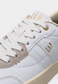 Baskets en cuir blanc avec lacets blancs, accent en daim beige, œillets dorés et logo "GAP" doré sur la languette et le côté.