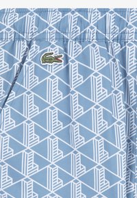 Ανοιχτό μπλε σορτς με γεωμετρικό λευκό σχέδιο και κεντημένο πράσινο λογότυπο κροκόδειλου. Κατασκευασμένο από ελαφρύ ύφασμα με ελαστική ζώνη μέσης.