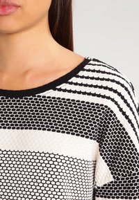 Czarnobiały sweter w paski wykonany na drutach, z szerokim dekoltem, fakturowanymi wzorami i różną szerokością pasków na materiale.