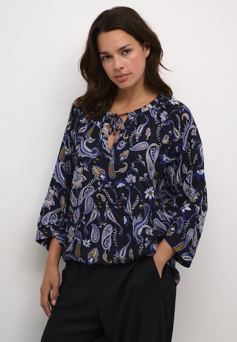 Blusa blu navy con motivi paisley e floreali. Presenta un collo con fiocco e maniche a tre quarti. Realizzata in un tessuto leggero e fluido.