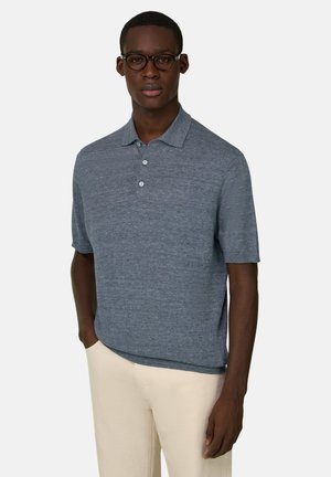 Mężczyzna w ciemnych okularach oprawkach, szarej koszulce polo z krótkim rękawem i guzikami oraz jasnobeżowych spodniach, stojący na tle jednolitego jasnego tła.