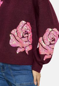 Maglione a coste bordeaux con grandi applicazioni di rose in paillettes rosa su entrambe le maniche. Tessuto morbido con un design a vestibilità rilassata.