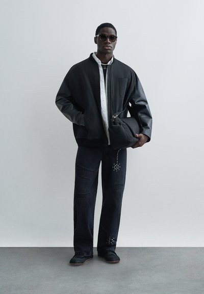 Geacă bomber neagră supradimensionată cu mâneci din piele, cămașă albă cu nasturi, blugi negri deteriorați cu accente cu ținte și încălțăminte neagră.