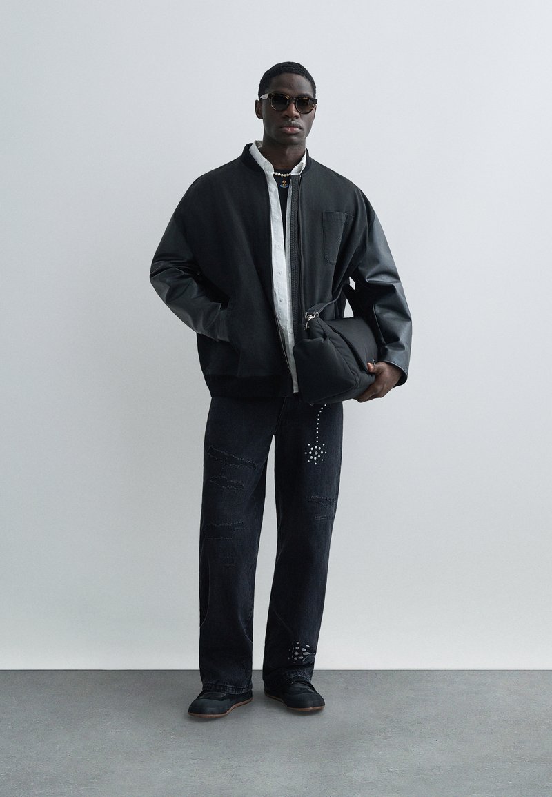 Bomber jacket oversize nero con maniche in pelle, camicia bianca con bottoni, jeans neri strappati con dettagli borchiati e calzature nere.