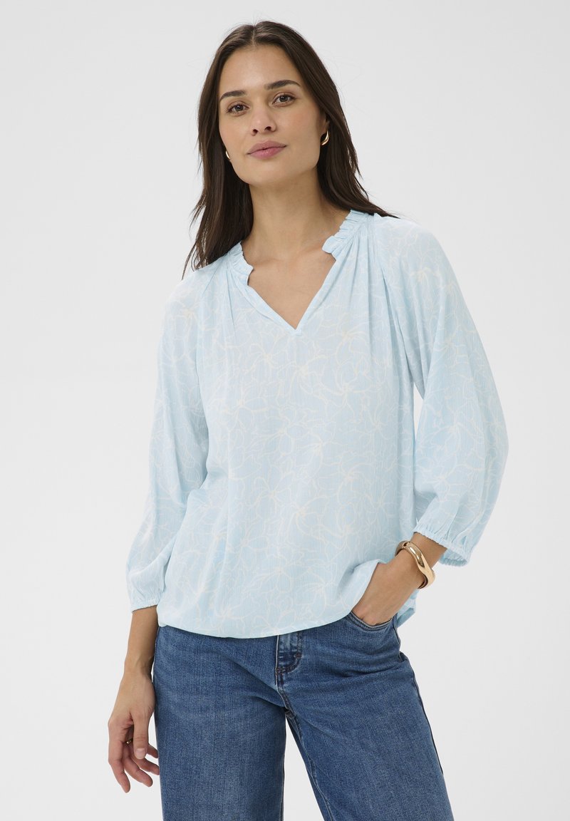 Femme portant un chemisier bleu clair à fleurs avec des manches trois-quarts et un jean bleu, debout avec une main dans la poche, sur un fond blanc.