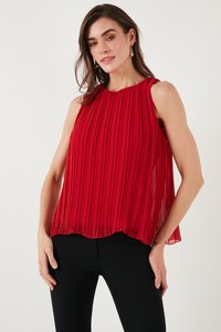 Woman wearing sleeveless red pleated blouse and black pants, standing with one hand on hip against plain background.