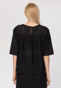 Blouse noire en coton avec motif à œillets, manches courtes et détail de décolleté en forme de clé. Elle présente une coupe ample et un design froncé.