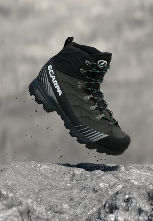 Zwart-grijze wandelschoen met blauwgroene veters, met het merk "SCARPA," zwevend boven rotsachtig terrein met vallende stofdeeltjes.