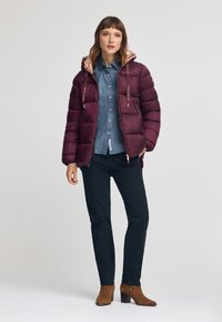 Mörkröd puffjacka med huva, roséguld accenter, dragkedja, framfickor och en kviltad design, bärs över en denimskjorta och mörka byxor.