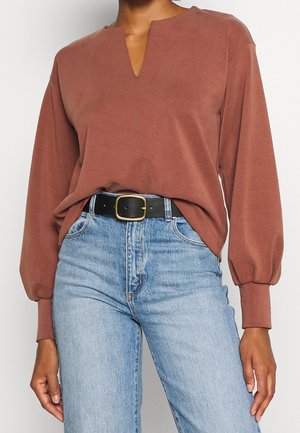 Frau trägt eine rostfarbene langärmelige Bluse mit Ausschnitt, die in hellblaue, hoch taillierte Jeans gesteckt ist, dazu ein schwarzer Gürtel mit goldener Schnalle.